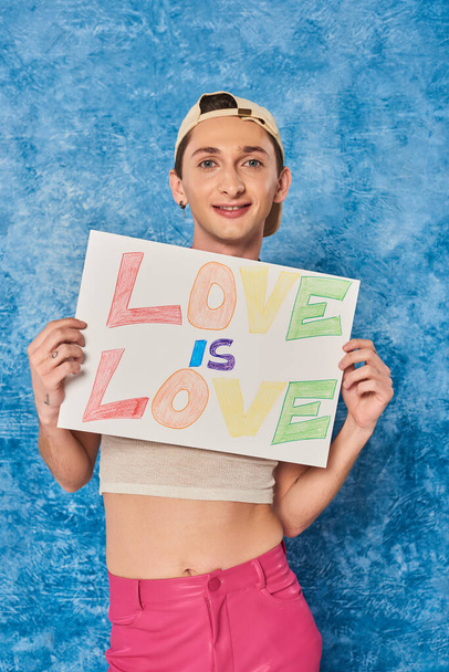 positiver schwuler Aktivist mit Baseballkappe und weißem Tanktop, der lächelt, während er ein Plakat mit Liebe in der Hand hält, sind Liebesworte während eines stolzen Monats auf fleckigem blauen Hintergrund  - Foto, Bild