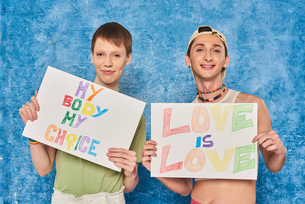 positieve gay activisten glimlachen terwijl het houden van plakkaten met liefde is liefde en mijn lichaam mijn keuze woorden tijdens trots maand op gevlekte blauwe achtergrond  - Foto, afbeelding