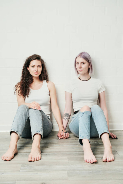 Portrait of content young Caucasian lesbians in jeans sitting on floor and holding hands - Fotó, kép