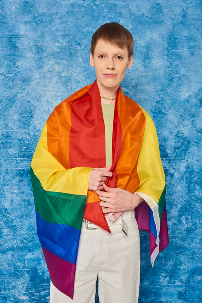 Portrait de jeune queer enveloppé dans un drapeau lgbt regardant la caméra et debout pendant la célébration du mois de la fierté sur fond bleu marbré - Photo, image