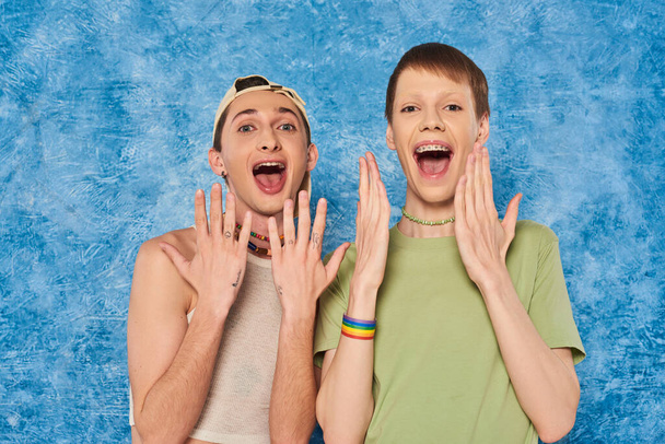 Šokovaní homosexuální přátelé v ležérní oblečení otevření úst a při pohledu na kameru během měsíce pýchy Igbt oslavy na skvrnité modré pozadí - Fotografie, Obrázek