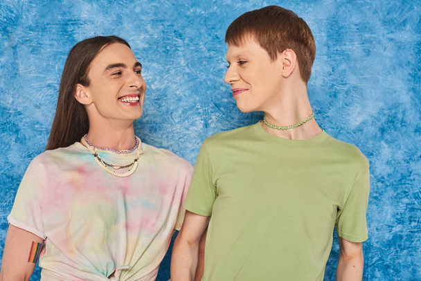 Portrait of cheerful queer friends in casual clothes looking at each other during lgbt pride month celebration on mottled textured blue background - Foto, imagen
