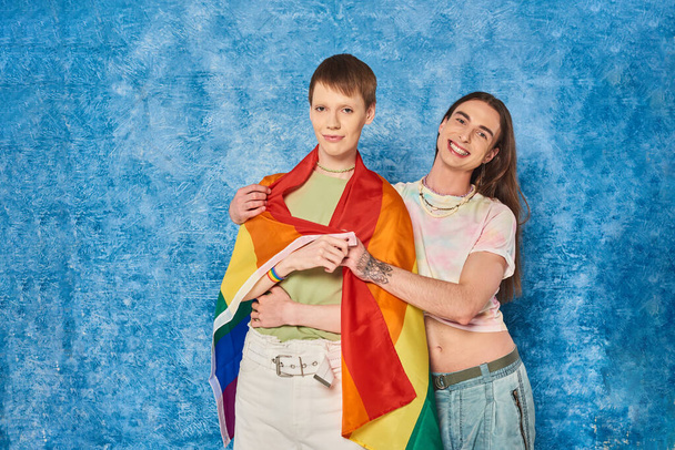 Sorrindo tatuado gay homem em roupas casuais abraçando jovem amigo com lgbt bandeira e olhando para a câmera durante o orgulho mês celebração no fundo azul manchado - Foto, Imagem