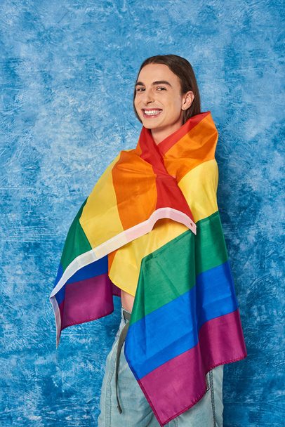 Positive long haired homosexual man looking and camera and wrapping in lgbt flag during pride community month celebration on mottled blue background - Valokuva, kuva