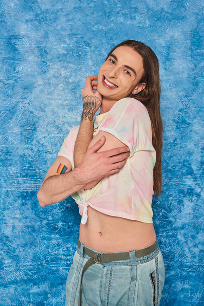 Carefree long haired and tattooed queer person smiling at camera and posing during lgbt month celebration on mottled and textured blue background - Photo, Image
