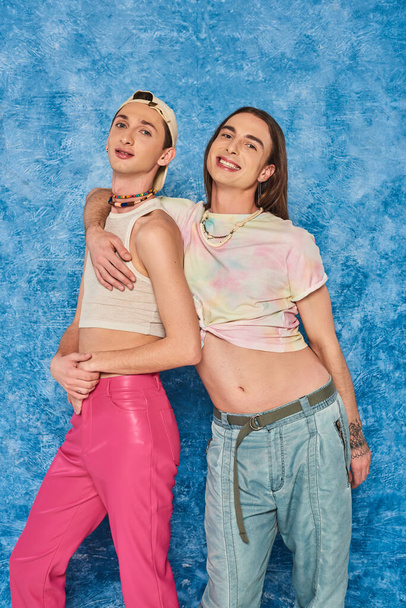 Portrait of smiling and stylish queer friends hugging and looking at camera together during lgbt pride month celebration on textured blue background - Foto, Imagem