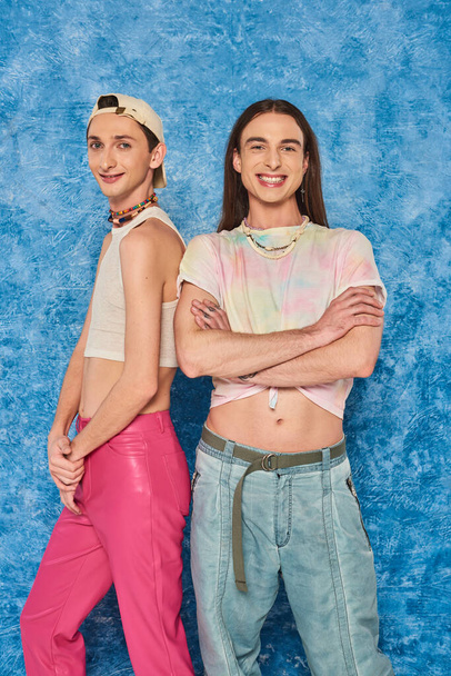 Stylish and young homosexual friends posing and smiling at camera together during lgbt community pride month celebration on textured blue background - Foto, immagini
