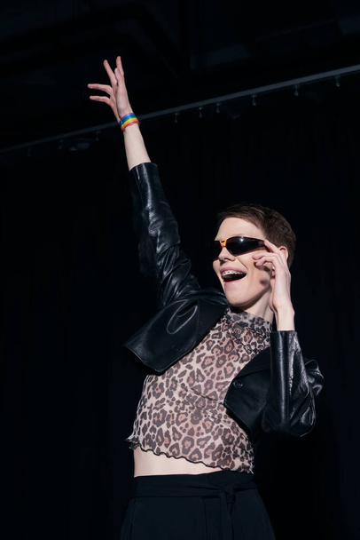 Carefree nonbinary person in stylish sunglasses, leather jacket and blouse with animal print celebrating pride month on black background  - Foto, Bild