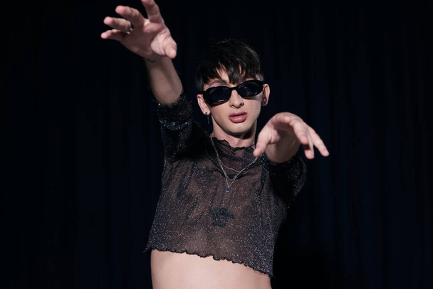 Fashionable and young homosexual man in sunglasses and shiny top outstretching hands at camera while celebrating lgbt pride month in isolated on black  - Foto, immagini