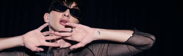 Retrato de joven, hombre gay tatuado y de moda en gafas de sol y la chispa posando durante lgbt orgullo mes partido aislado en negro, pancarta  - Foto, Imagen