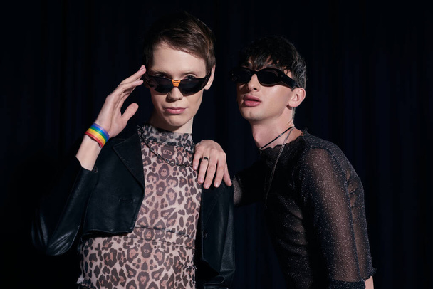 Portrait of fashionable queer persons in sunglasses and party outfits posing and looking at camera during lgbt pride month celebration isolated on black  - Фото, зображення