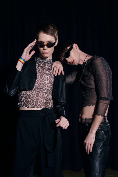 Trendy and young gay friends in sunglasses and party outfits posing and looking at camera during lgbt pride month celebration isolated on black - 写真・画像