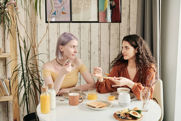 Happy young lesbians sitting at dining table and chatting while eating toasts with jam at home - Photo, Image
