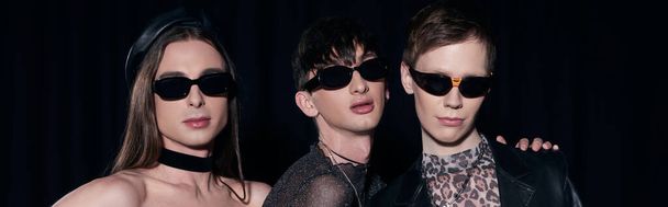 Portrait of fashionable homosexual couple in party clothes and sunglasses posing during celebration of lgbt pride month isolated on black, banner  - Фото, изображение