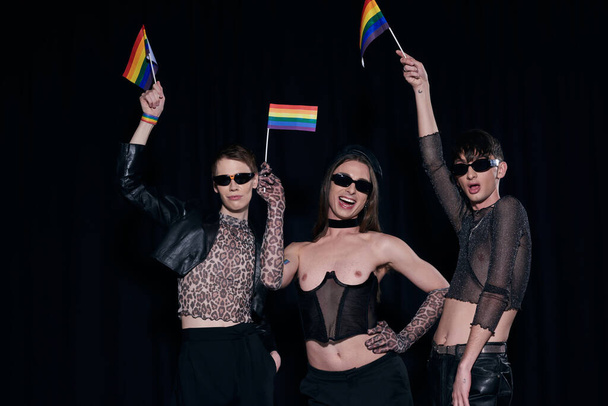 Ξέγνοιαστοι και μοντέρνοι ομοφυλόφιλοι φίλοι με γυαλιά ηλίου και ρούχα για πάρτι κρατώντας σημαίες ουράνιου τόξου κατά τη διάρκεια εορτασμού μήνα υπερηφάνειας  - Φωτογραφία, εικόνα