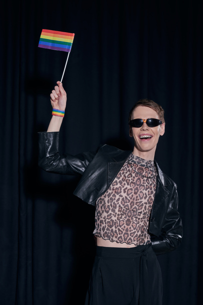 Cheerful and fashionable nonbinary person in leather jacket and top with animal print holding lgbt flag during pride month celebration on black background  - Foto, imagen