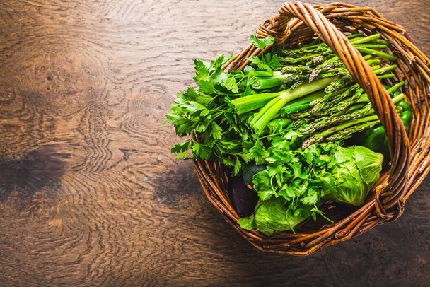 Légumes verts frais et herbes dans un panier en osier, asperges, laitue, courgettes avec persil et coriandre sur table en bois - Photo, image