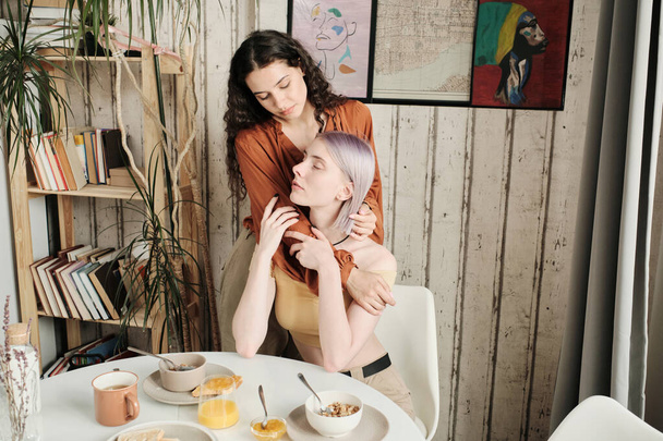 Serene young woman with closed eyes sitting at dining table with breakfast and enjoying hug of girlfriend - Foto, Bild