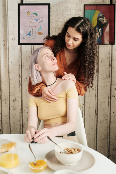 Wavy-haired woman embracing girlfriend from back and wishing good day to her during breakfast - Zdjęcie, obraz