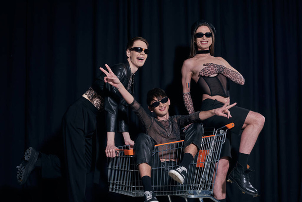 Smiling and stylish group of homosexual friends in sunglasses having fun with friend in shopping cart during pride month celebration on black background  - Valokuva, kuva