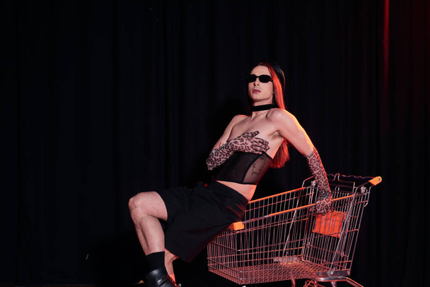 Fashionable homosexual man in sunglasses and sexy corset sitting on shopping cart during party and lgbt pride month celebration on black background  - Photo, Image