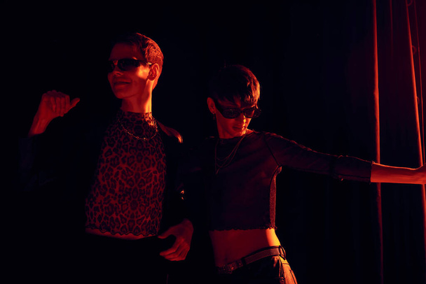 Fashionable nonbinary friends in party outfits and sunglasses dancing together while celebrating lgbt pride month on black background with red lighting  - Photo, Image