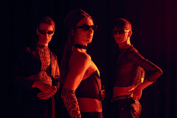 Grupo de la comunidad homosexual de moda en gafas de sol y trajes de fiesta posando juntos mientras se celebra lgbt mes aislado en negro con iluminación roja  - Foto, Imagen