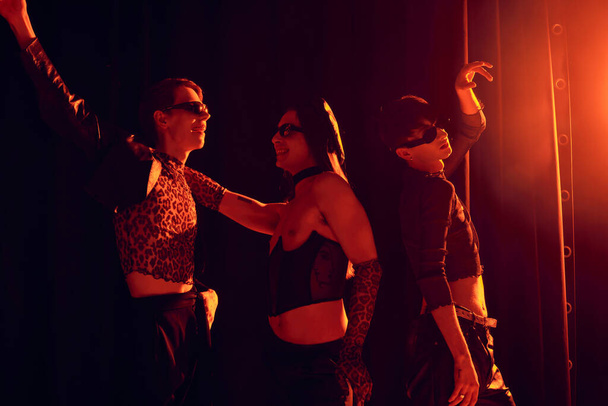 Smiling and fashionable homosexual people in sunglasses dancing during party and lgbt pride month celebration on black background with red lighting  - Photo, Image