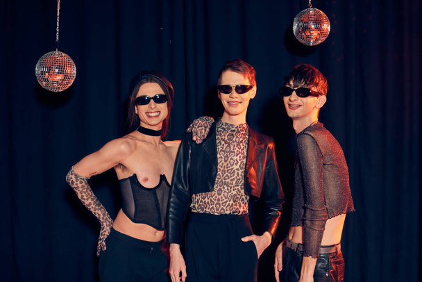 Cheerful and fashionable gay community in party outfits and sunglasses posing together near disco balls during pride month celebration on black background  - Zdjęcie, obraz