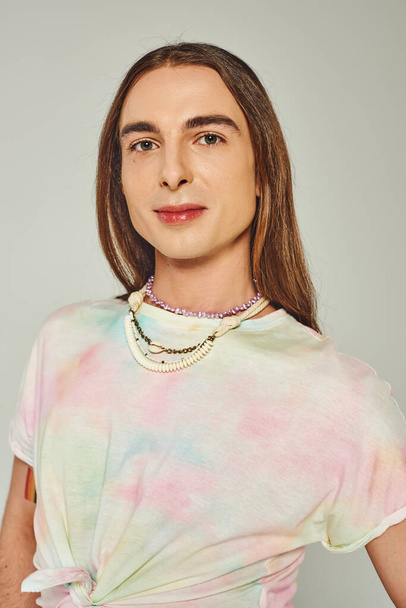 Portrait of young and long haired homosexual man in tie dye t-shirt looking at camera isolated on grey, happy pride community month celebration concept  - Foto, Bild