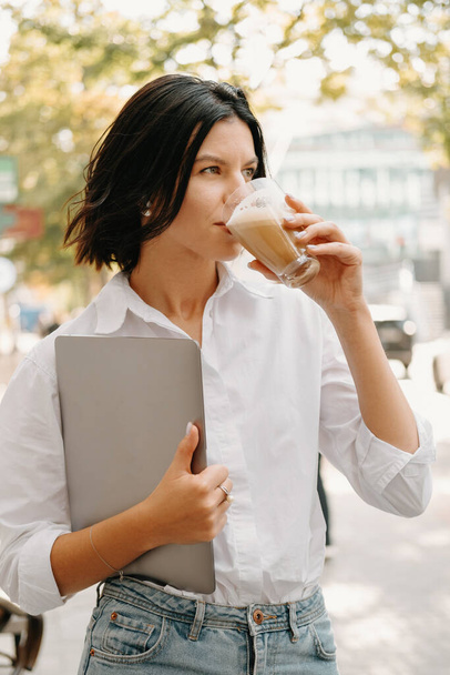 Giovane donna bussines sta camminando all'aperto mentre tiene il computer portatile e beve un latte macchiato. - Foto, immagini
