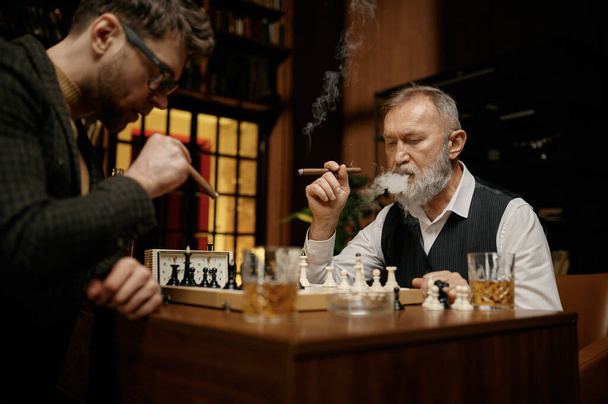 Rodzina inteligentnych ludzi grająca w szachy, paląca cygara i pijąca whisky. Starsi i młodsi mężczyźni siedzący przy stole i patrzący na szachownicę. - Zdjęcie, obraz