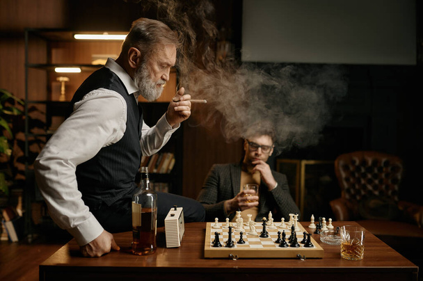 Two chess players taking break for smoking cigar and planning strategy of game. Selective focus on senior man looking at chessboard - Photo, Image