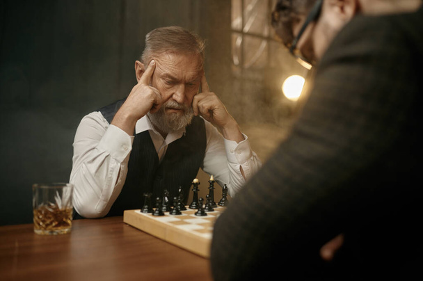 Старший шахматист трогает храмы, чувствуя себя потерянным в мыслях, планируя тактический ход для борьбы с молодым противником - Фото, изображение