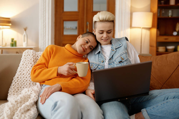 Happy loving lgbt woman couple watching video online using laptop while sitting and resting on sofa couch in cozy home living room - Foto, afbeelding