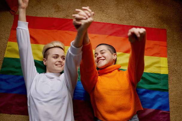 Vue de dessus sur heureux jeune couple lesbien couché au-dessus du drapeau arc-en-ciel lgtb avec les mains levées. Concept communautaire LGBTQIA - Photo, image