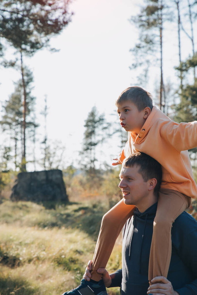 Papá e hijo en un paseo por el bosque en un clima soleado. De ocio familiar. El padre lleva a su hijo sobre sus hombros. Vista frontal - Foto, imagen