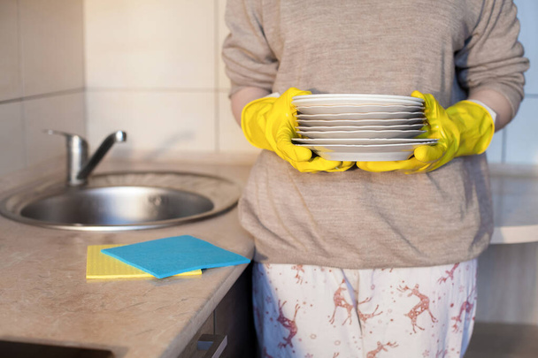 Девушка в жёлтых резиновых перчатках держит чистые тарелки в руках на кухне. Сюжет о домашней работе, чистоте и повседневной деятельности. - Фото, изображение