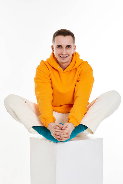 young toothy guy in orange sweatshirt sitting on white cube on white background. Full length - Foto, afbeelding