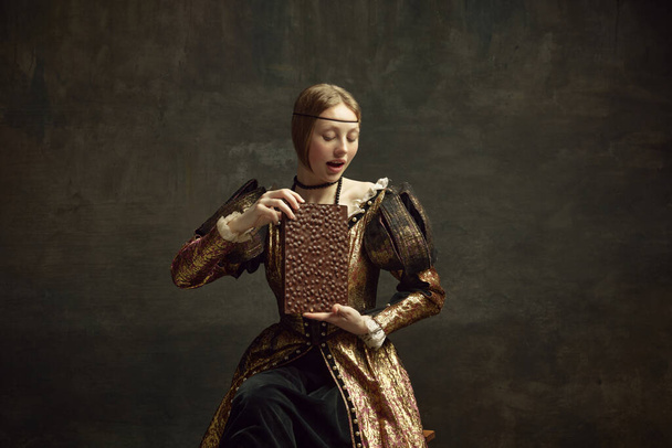Portrait of young girl, medieval royal person, pretty princess in vintage dress holding giant chocolate bar over dark green background. Concept of history, renaissance art remake, comparison of eras - Foto, Bild