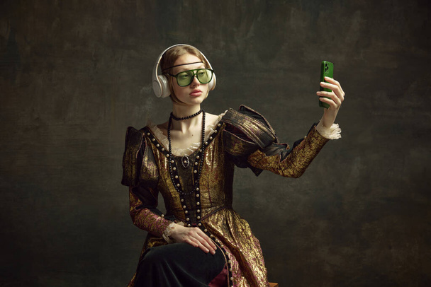 Portrait of young beautiful girl in vintage dress, trendy sunglasses and headphones, taking selfie with phone against dark green background. Concept of history, renaissance art, comparison of eras - Photo, image