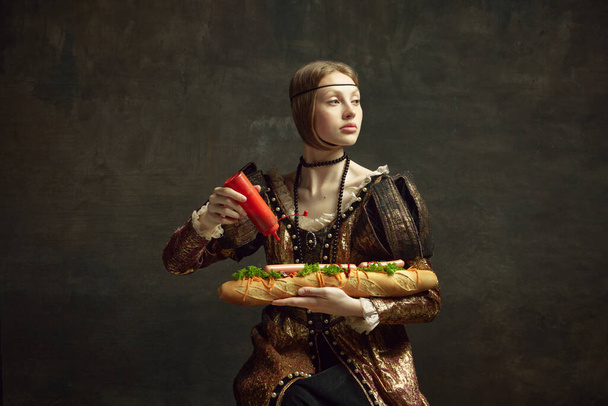 Portrait of young girl, queen, princess in vintage costume putting ketchup on giant sandwich baguette on dark green background. Concept of history, renaissance art, comparison of eras, health and food - 写真・画像