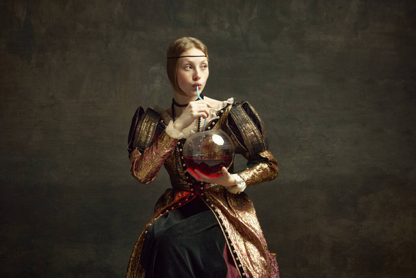 Portrait of beautiful young royal person, girl in vintage dress drinking wine from giant glass against dark green background. Party time. Concept of history, renaissance art remake, comparison of eras - Foto, Imagem