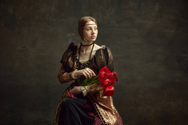 Portrait of beautiful young girl, royal person, queen in elegant vintage dress posing with flowers, tulips against dark green background. Concept of history, renaissance art remake, comparison of eras - Photo, Image
