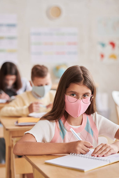 Retrato vertical da menina usando máscara na escola e olhando para a câmera enquanto estava sentado na mesa em sala de aula - Foto, Imagem
