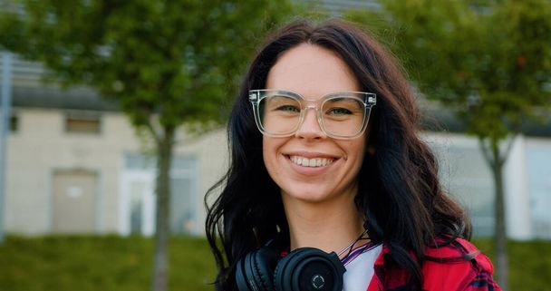 Portrait of lovely happy smiling young woman with brown wavy hair in glasses and with headphones which standing in front of camera near city building with green plantings near it - Φωτογραφία, εικόνα