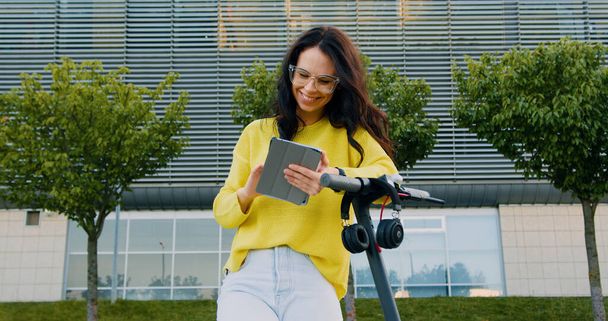 Pretty joyful carefree young brown-haired woman in stylish clothes leaning on her e-scooter and using tablet computer on the background of modern big building with green trees near it - Фото, зображення
