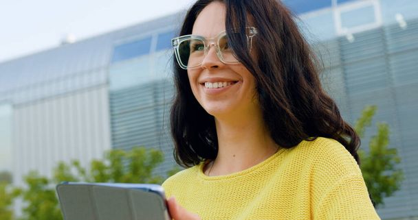 Lovely happy smiling stylish young brunette in glasses using her portable device on the background of big panoramic building at daytime - 写真・画像