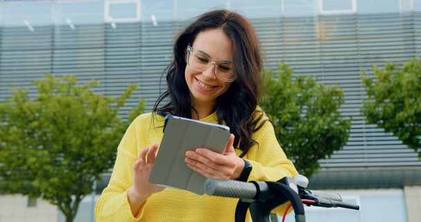 Cute friendly in high spirits young smiling brunette in stylish clothes and glasses using her tablet pc while standing near her electric scooter on the big modern building background at daytime - Valokuva, kuva