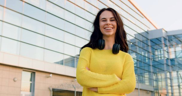 Портрет красивой улыбающейся счастливой беззаботной стильной юной брюнетки, которая позирует на камеру со скрещенными руками рядом с панорамным стеклянным зданием днем - Фото, изображение